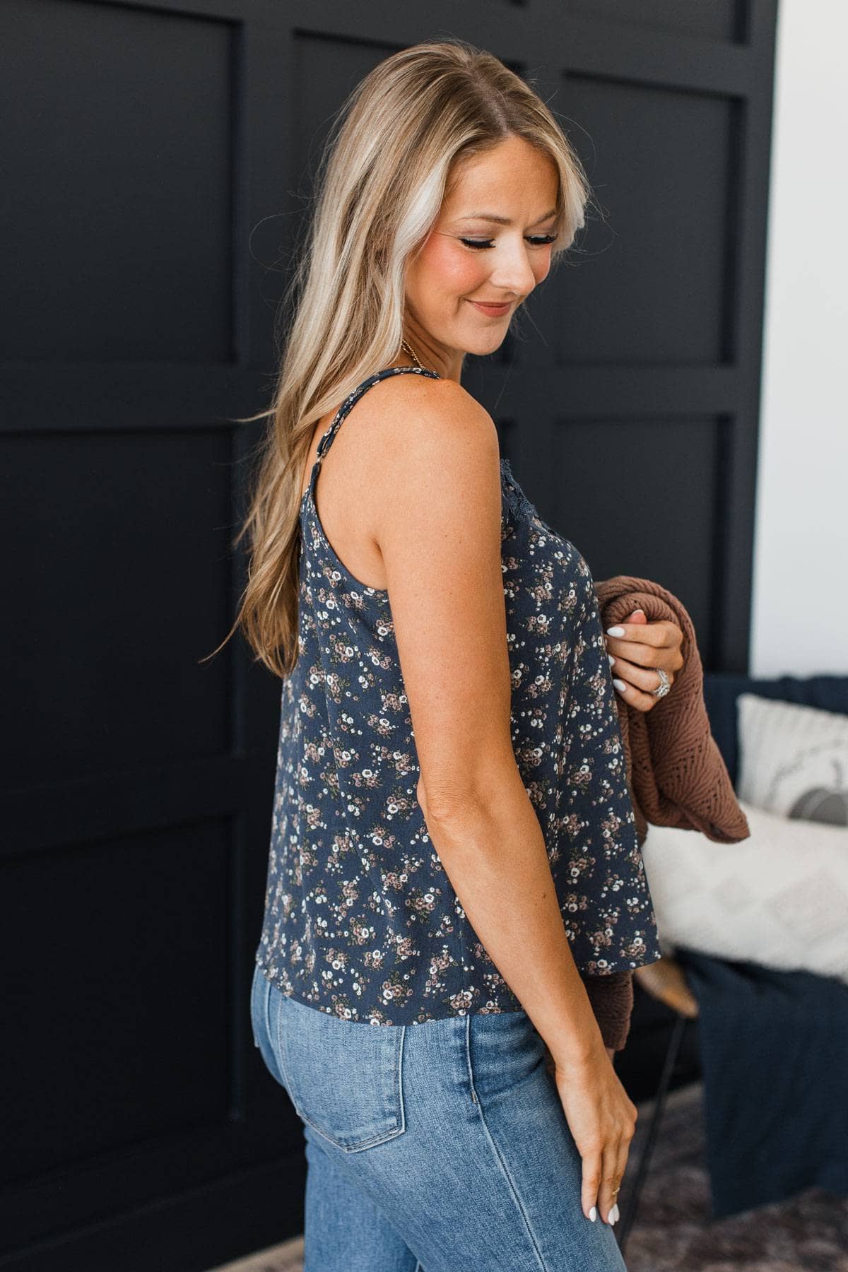 A woman wearing a navy blue floral tank top and blue jeans, holding a brown cardigan in a bedroom setting with a dark paneled wall behind her.