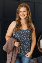 A smiling woman with long wavy light brown hair stands indoors against a dark paneled wall, wearing a sleeveless navy floral camisole top and blue jeans while holding a brown knit cardigan.