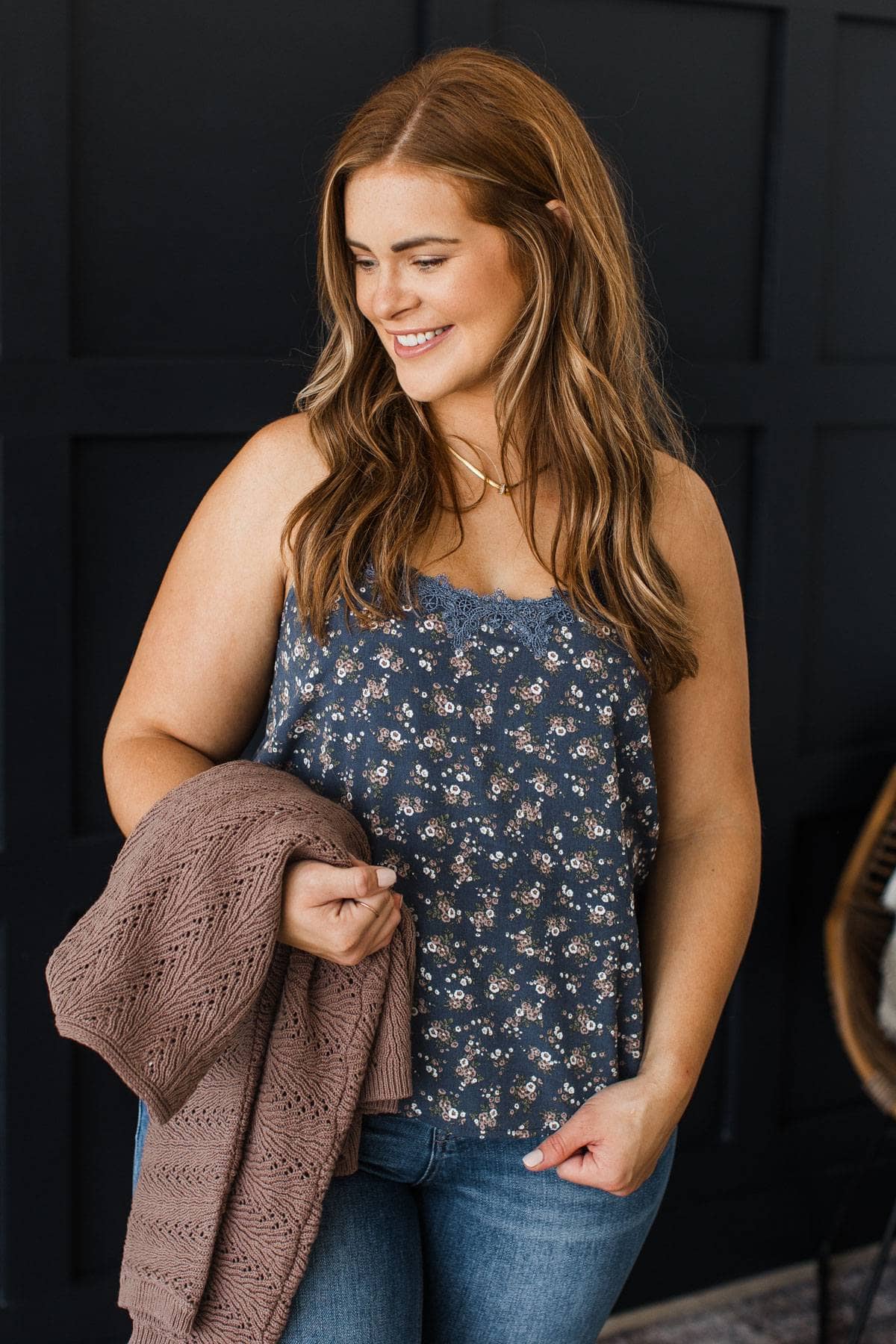 A woman with long light brown hair is shown from the waist up, wearing a sleeveless navy blue floral camisole top and blue jeans, while holding a brown knit blanket against a dark paneled wall.