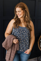A woman with long light brown hair is shown from the waist up, wearing a sleeveless navy blue floral camisole top and blue jeans, while holding a brown knit blanket against a dark paneled wall.